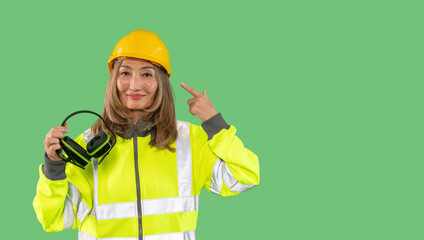 Woman wearing safety gear and hard hat in a green background while pointing to her ear protection