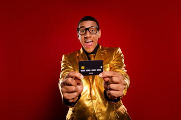 Charismatic man in a golden suit joyfully displaying a credit card against a vibrant red background