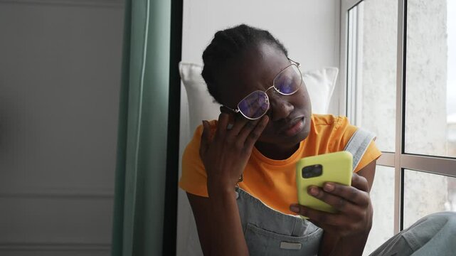 Young black woman with glasses experiencing eye strain while sitting near window, rubbing tired eyes after prolonged smartphone use and feeling digital device-related discomfort