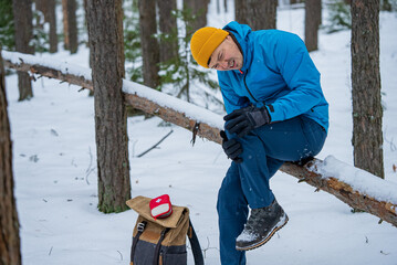 Male hiker experiencing severe knee pain while sitting on fallen tree trunk in snowy forest, with first aid kit nearby during winter hiking trip