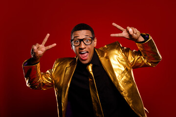 Charismatic young man in golden jacket poses with peace signs against a vivid red backdrop,...
