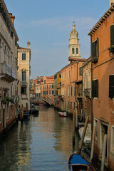 A tranquil canal winds through Venice, Italy, reflecting the vibrant hues of historic buildings and a distant church tower. This is a classic Venetian view that showcases the beauty of this city.