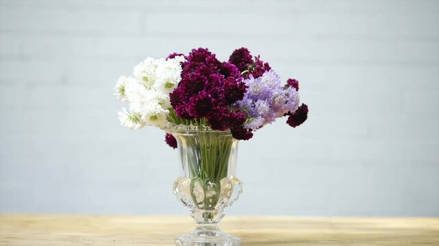 A vase filled with purple and white flowers sits on a wooden table. The flowers are arranged in a way that creates a sense of balance and harmony. The vase itself is clear