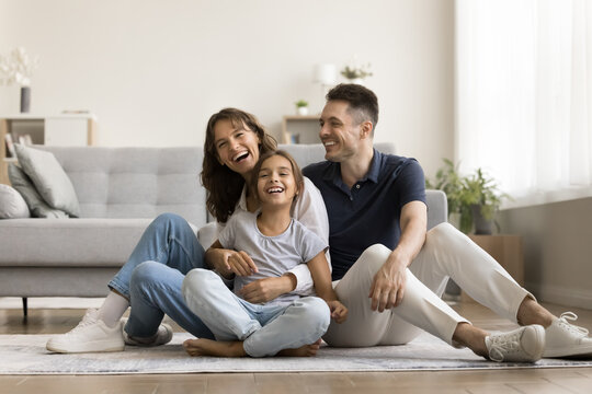 Happy Hispanic family with cheery cute daughter relaxing seated on warm floor in living room, laughing, looking carefree, feeling unconditional love, relish untroubled weekend together at modern home