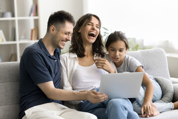 Married couple and little daughter sitting on couch using computer watching funny comedy movie, laughing, enjoy online content or e-shopping, resting on carefree leisure on internet. Modern technology