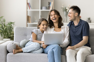 Young laughing parents with preschool daughter resting on sofa watching family comedy movie through...