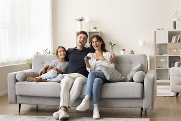 Family members, young parents and cute little daughter sit on sofa at home, switch channels, watching comedy movie or educational program, enjoy shared activity, togetherness and quality time together