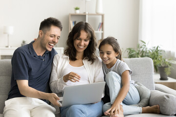 Fototapeta premium Young parents with their little daughter watching online movies, comedy program or cartoons and laughing, sitting, resting on sofa with modern wireless computer, spend free time on internet. Tech, fun