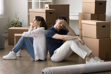 Tired frustrated married couple sitting back-to-back on floor surrounded by cardboard moving boxes,...