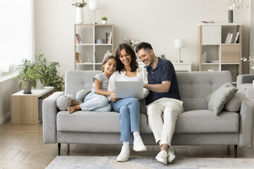 Married couple and little preschooler daughter sitting on couch using modern laptop, watching...