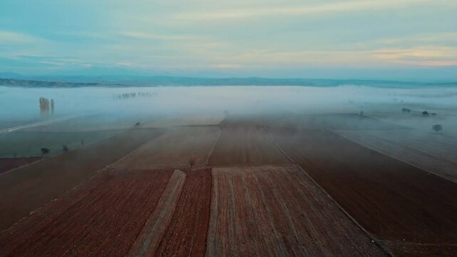 Fog rolling over a countryside road on a mountain slope. Aerial view of fog covering agricultural land and vacant areas. Mystical and misty mountain landscape in serene, foggy weather.