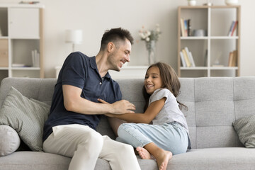 Happy little daughter sits on sofa with daddy, laughing, playing together, tickling each other,...