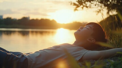 Person laying on the grass next to a body of water during sunset, enjoying the serene atmosphere.