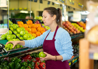Positive middle-aged saleswoman setting fresh apples on food stall in grocery store
