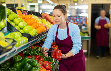 Positive middle-aged saleswoman setting out big peppers on food stall in grocery store