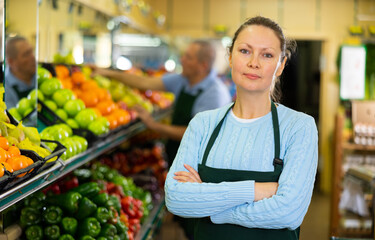 Smiling middle-aged shop assistant posing in front of counter in greengrocery with big stock
