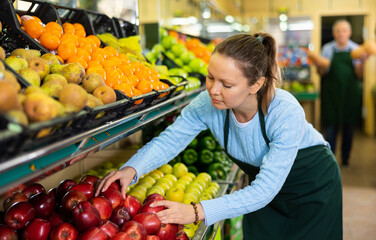 Positive middle-aged saleswoman setting fresh apples on food stall in grocery store