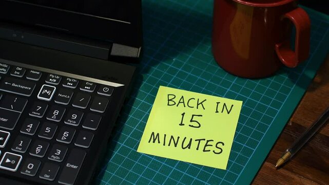 Man&rsquo;s hand sticking yellow Back in 15 minutes note message to office desk mood lighting copy space