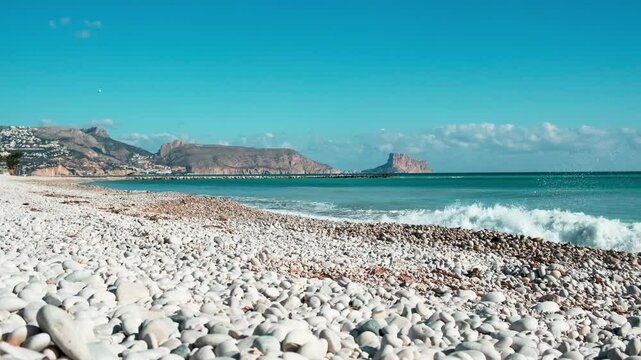 Beautiful white pebble beach in Altea and rolling Mediterranean Sea. View from Altea to turquoise Mediterranean sea with waves, Calpe city and Rock of Ifach. Altea, Alicante province, Spain
