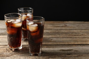Refreshing cola with ice cubes in glasses on wooden table against black background, closeup. Space for text