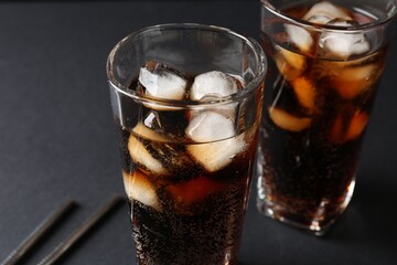 Refreshing cola with ice cubes in glasses and drinking straws on black table, closeup