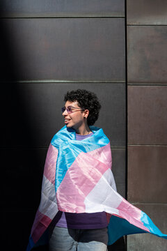 LGBTQ+ person smiles wrapped in transgender pride flag outdoors