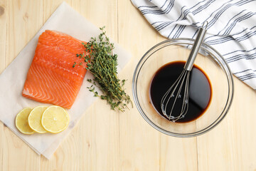 Soy sauce in bowl, whisk, salmon fillet, thyme and lemon slices on wooden table, flat lay