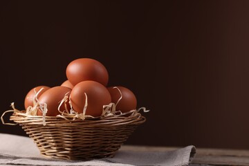 Wicker basket with fresh eggs on wooden table against brown background, closeup. Space for text