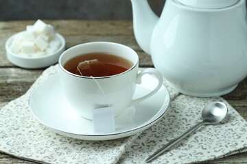 Tea bag in cup with hot drink, teapot and spoon on wooden table, closeup