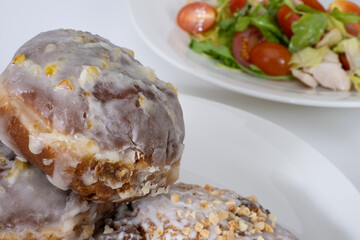 A close up of glazed donuts with crushed nuts in the foreground, paired with a fresh salad of cherry tomatoes, lettuce, and chicken on a white plate.