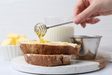 Woman pouring honey onto slices of bread with butter at white table, closeup