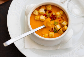 Spanish cold soup gazpacho in bowl with tomatoes, cucumber, baked breads and onion on table in restaurant