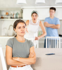 Obraz premium Sad offended young woman sitting at kitchen table during argument with dissatisfied husband and mother-in-law standing behind on blurred background. Concept of family fights