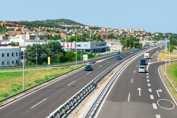 Obraz premium Modern highway with multiple lanes, surrounded by green landscapes and urban buildings in the background, under a clear blue sky. Ideal for transportation, infrastructure or travel-related themes
