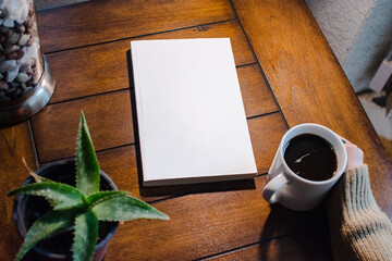 Photo of a blank book on a wooden table next to a plant and a mug of coffee held by a female hand