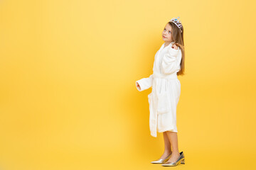 A preteen girl stands confidently against a vivid yellow background. She wears a fluffy white robe and a sparkling crown, showcasing her stylish personality and playful spirit, copy space