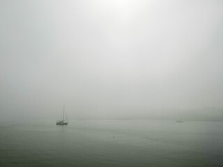 Fototapeta premium A tranquil scene of a sailboat drifting on calm waters, surrounded by dense fog that obscures the horizon. The minimalist composition and soft tones evoke feelings of solitude, serenity, and mystery