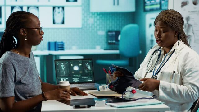 Black medic practitioner takes blood pressure with a blood pressure monitor, does a consultation with female patient using modern advanced tools for the checkup routine. Camera A.