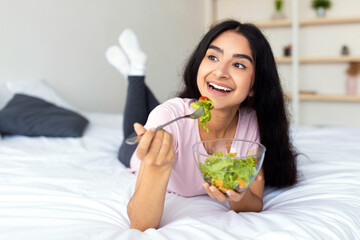 Weight loss diet concept. Pretty Indian woman eating fresh vegetable salad on bed at home, full...
