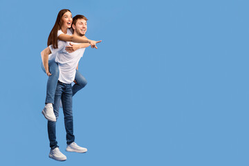 Surprised couple pointing finger aside at copy space isolated over purple studio wall, boy giving his girl piggyback ride