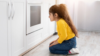 Cute little girl sitting near oven in kitchen and looking inside through glass, curious impatient...