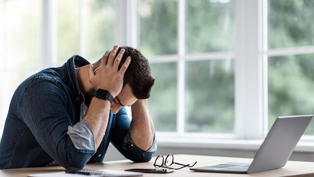 Tired stressed young man suffering from headache attack after computer work, feeling fatigue at home, overworked. Exhausted unhappy guy touches temples to relieve pain from migraine high pressure