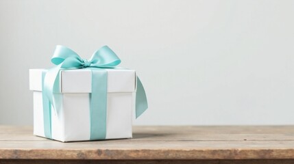 A single white gift box adorned with a delicate light blue ribbon sits on a rustic wooden surface against a neutral backdrop