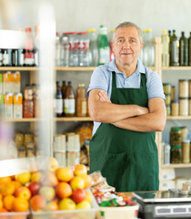 Mature man seller in apron posing at counter in grocery store