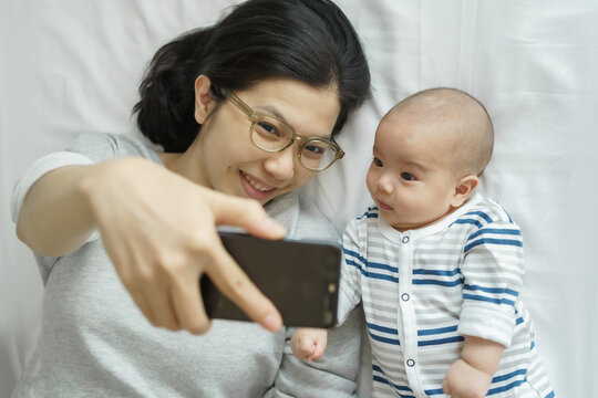 Asian Mother taking a selfie with baby lying on bed. Overhead shot of bonding moment. Parenting and technology concept - Powered by Adobe