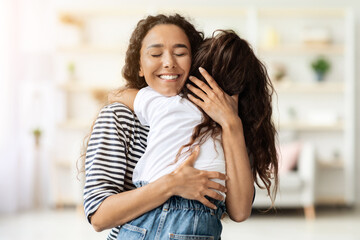 Fototapeta premium Happy middle-eastern woman hugging her little curly girl kid while spending time together at home, closeup portrait, blurred background. Mother and daughter, family, motherhood concept