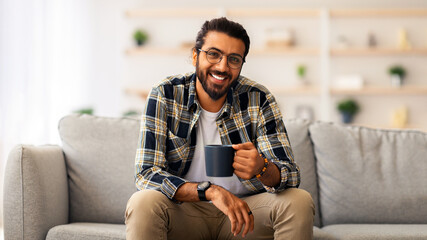 Handsome bearded millennial arab man resting at home, sitting on couch in cozy living room,...