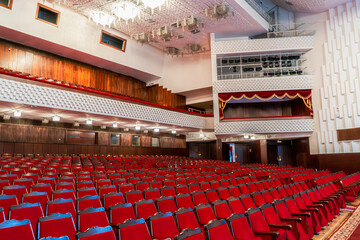 Naklejka premium This image showcases a grand theater interior featuring rows of red velvet seats facing the stage. A blend of modern and classical elements is visible in the rich wood paneling, ornate balconies
