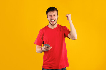Yes. Portrait of happy emotional young man using smartphone making winner gesture isolated over blue studio wall background. Excited guy celebrating win, shaking clenched fist, looking at camera