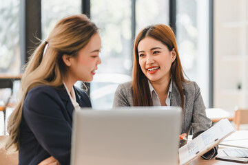 Two businesswomen in formal attire discussing work at desk with laptop and documents, smiling and engaging in bright office environment